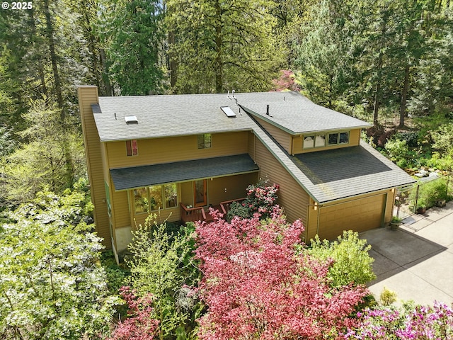 view of front of property featuring roof with shingles, an attached garage, a view of trees, a chimney, and driveway