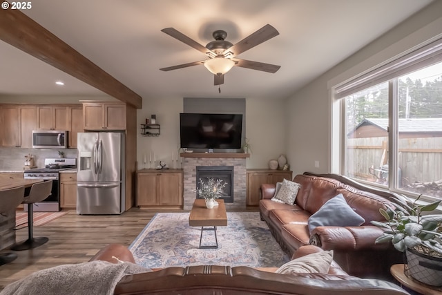 living room with ceiling fan, light wood-type flooring, beamed ceiling, and a stone fireplace