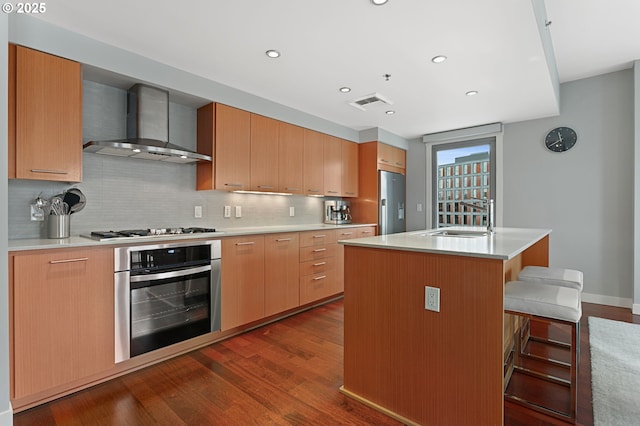 kitchen featuring wall chimney exhaust hood, a kitchen bar, sink, a center island with sink, and appliances with stainless steel finishes