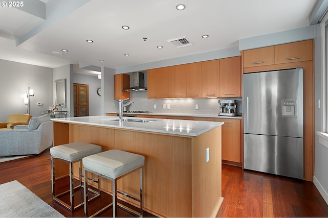 kitchen featuring dark wood-type flooring, wall chimney exhaust hood, stainless steel fridge, and a kitchen island with sink