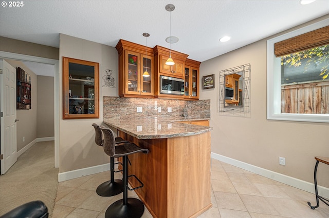 kitchen featuring stainless steel microwave, brown cabinetry, light stone counters, a peninsula, and glass insert cabinets