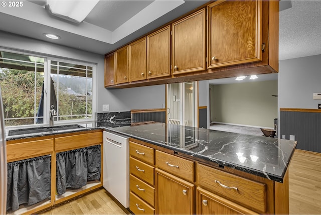 kitchen featuring dark stone countertops, light wood finished floors, white dishwasher, a sink, and brown cabinets