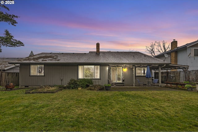 back of property at dusk featuring a patio area, a lawn, a shingled roof, and fence