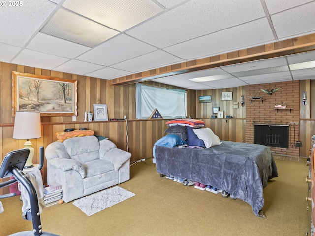 carpeted bedroom featuring a brick fireplace, wooden walls, and a drop ceiling