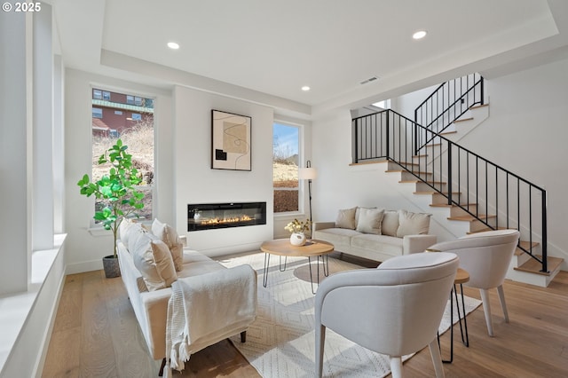 living area with a raised ceiling, a glass covered fireplace, wood finished floors, and recessed lighting