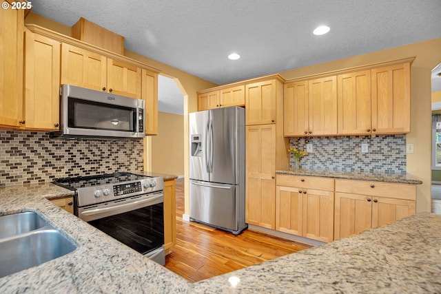 kitchen featuring light stone counters, light brown cabinets, stainless steel appliances, light wood-type flooring, and decorative backsplash