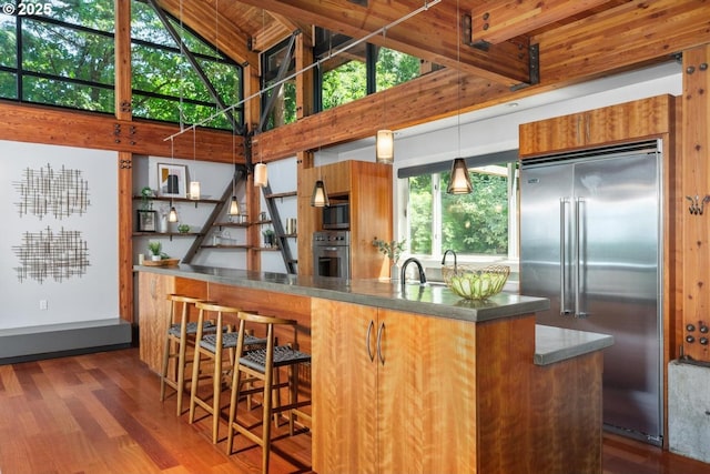 4183 SW Greenleaf Dr, Portland, OR 97221 - kitchen with stainless steel appliances, high vaulted ceiling, brown cabinetry, dark wood-style flooring, and decorative light fixtures