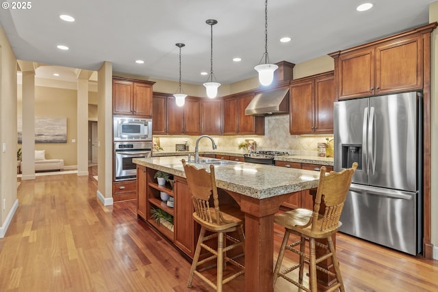 kitchen with sink, a center island with sink, stainless steel appliances, light stone countertops, and wall chimney range hood