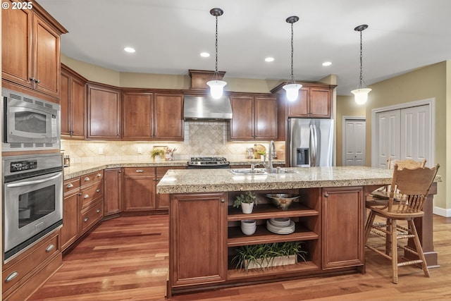 kitchen with wall chimney exhaust hood, sink, hanging light fixtures, a center island with sink, and appliances with stainless steel finishes