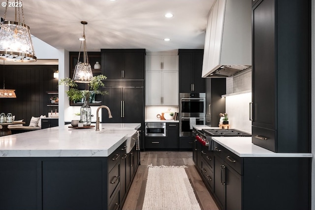 kitchen with light stone counters, a kitchen island with sink, stainless steel appliances, light wood-type flooring, and pendant lighting