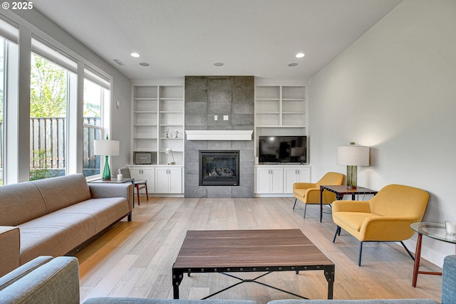 living room featuring light wood finished floors, a textured ceiling, a tiled fireplace, built in features, and visible vents