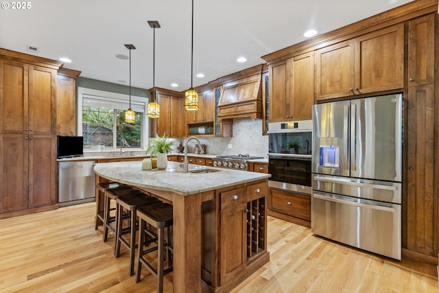 kitchen with a sink, light wood-style floors, appliances with stainless steel finishes, brown cabinetry, and custom exhaust hood