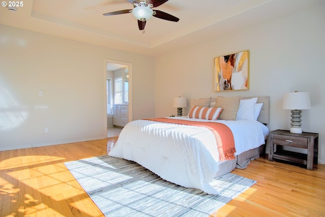 bedroom with a tray ceiling, ceiling fan, ensuite bath, and light wood finished floors