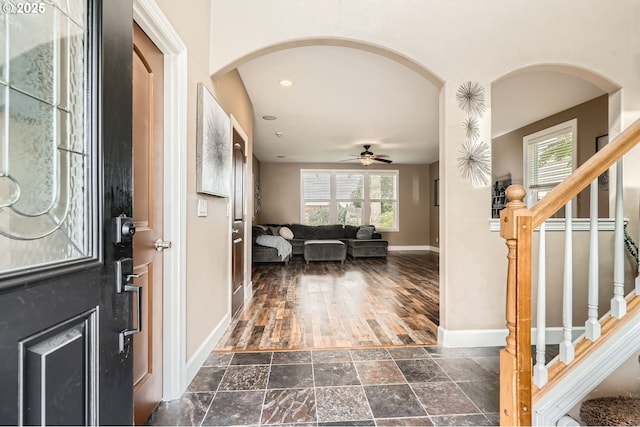 foyer featuring stairway, a ceiling fan, stone finish floor, baseboards, and arched walkways