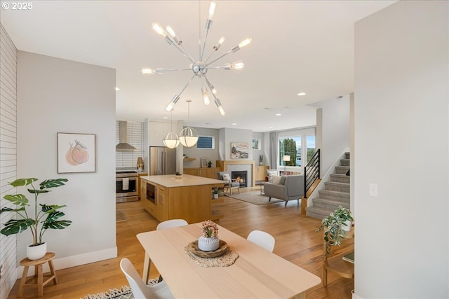 dining area with a warm lit fireplace, a notable chandelier, light wood-style flooring, stairs, and recessed lighting