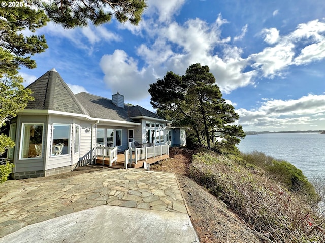 rear view of house featuring a shingled roof, a deck with water view, a patio, and a chimney