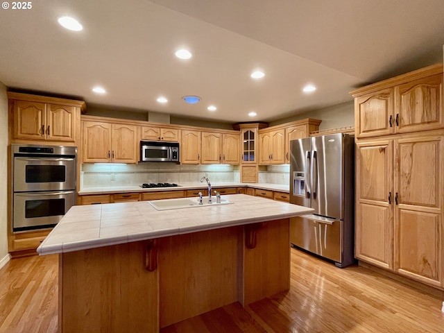 kitchen with tile counters, light wood-style flooring, a kitchen island with sink, and appliances with stainless steel finishes