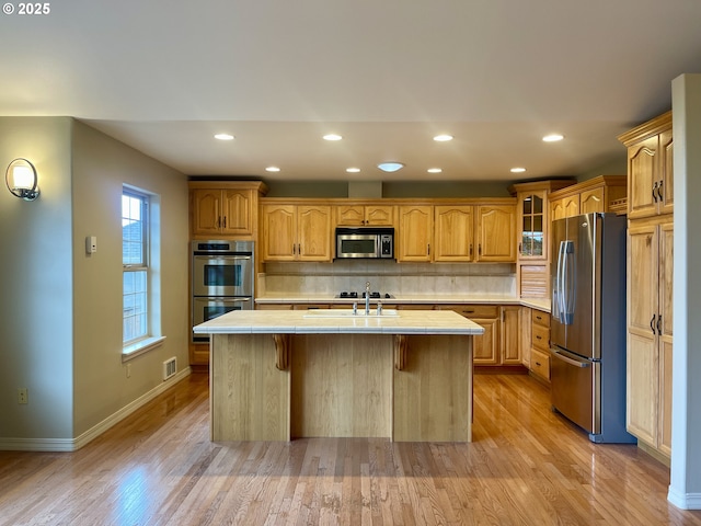 2180 Hayes St, North Bend, OR 97459 - kitchen featuring stainless steel appliances, tile countertops, light wood-type flooring, a kitchen island with sink, and tasteful backsplash
