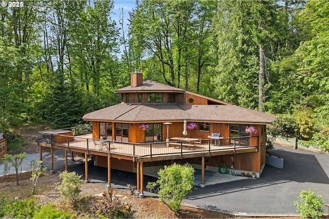 rear view of property featuring a wooden deck, a chimney, roof with shingles, and a view of trees