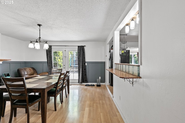 dining room with a chandelier, a wainscoted wall, light wood-style flooring, a textured ceiling, and french doors