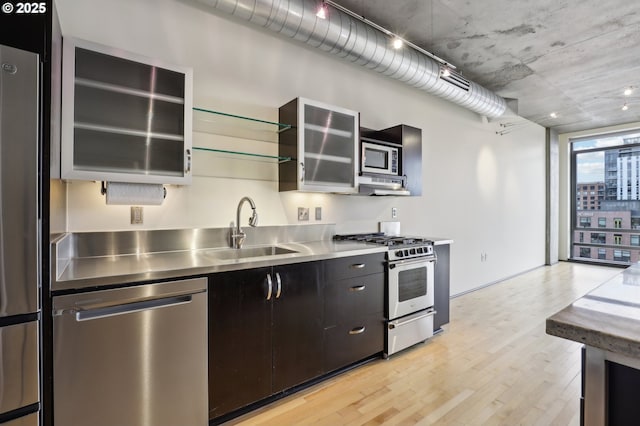 kitchen with glass insert cabinets, stainless steel countertops, light wood-style flooring, stainless steel appliances, and a sink