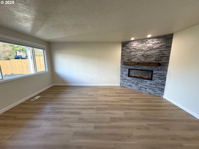 unfurnished living room with visible vents, a stone fireplace, a textured ceiling, wood finished floors, and baseboards