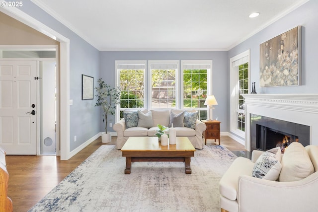 living area with wood finished floors, baseboards, crown molding, a warm lit fireplace, and recessed lighting
