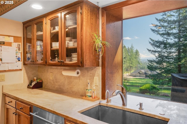 kitchen featuring brown cabinetry, a sink, light countertops, glass insert cabinets, and tasteful backsplash