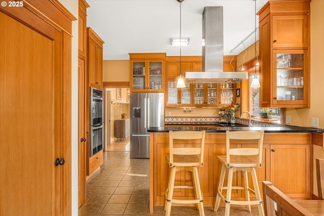 kitchen featuring dark countertops, glass insert cabinets, island range hood, brown cabinets, and appliances with stainless steel finishes