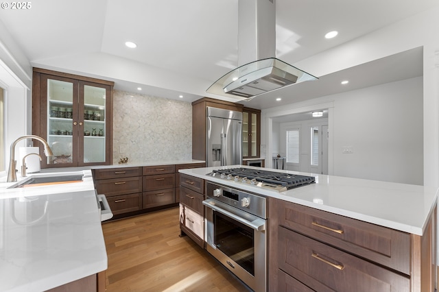 kitchen featuring a sink, appliances with stainless steel finishes, light wood-type flooring, island exhaust hood, and glass insert cabinets