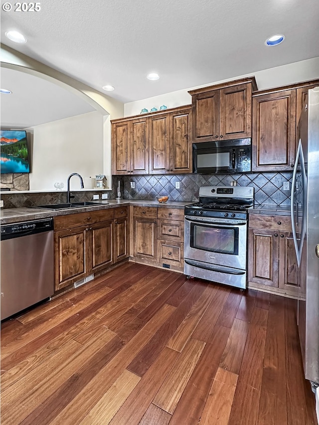 kitchen featuring dark wood finished floors, arched walkways, a sink, decorative backsplash, and appliances with stainless steel finishes