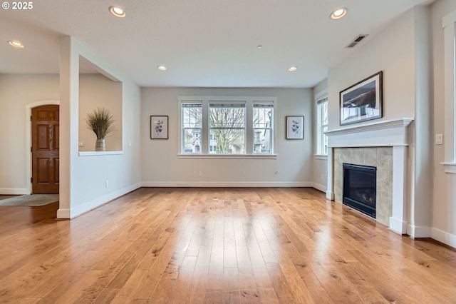 unfurnished living room featuring a tiled fireplace, light wood-style flooring, baseboards, recessed lighting, and arched walkways