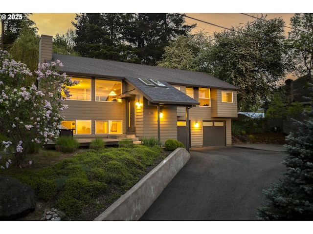 raised ranch featuring an attached garage, a shingled roof, driveway, and a chimney