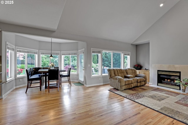 living room with healthy amount of natural light, light wood-type flooring, baseboards, a glass covered fireplace, and high vaulted ceiling