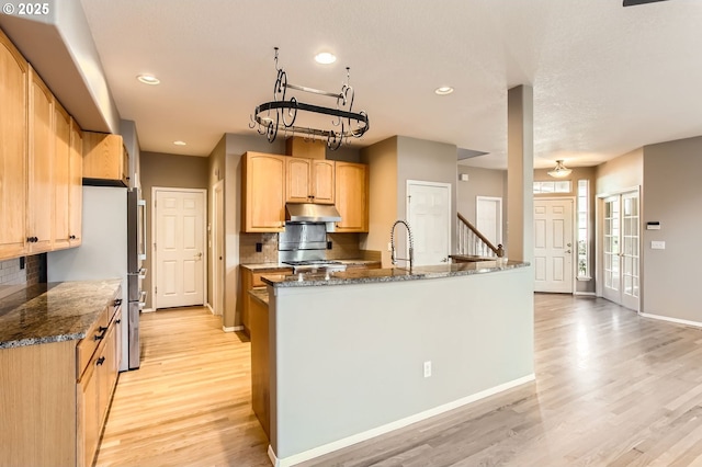 kitchen featuring light wood finished floors, baseboards, dark stone countertops, under cabinet range hood, and light brown cabinets