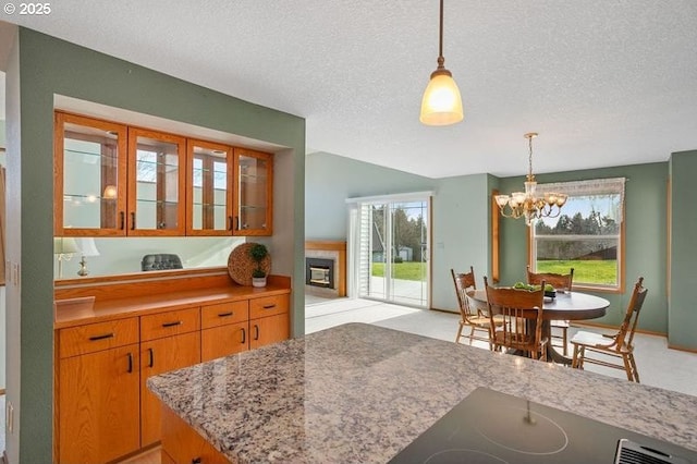 kitchen with a textured ceiling, glass insert cabinets, brown cabinetry, a tiled fireplace, and decorative light fixtures