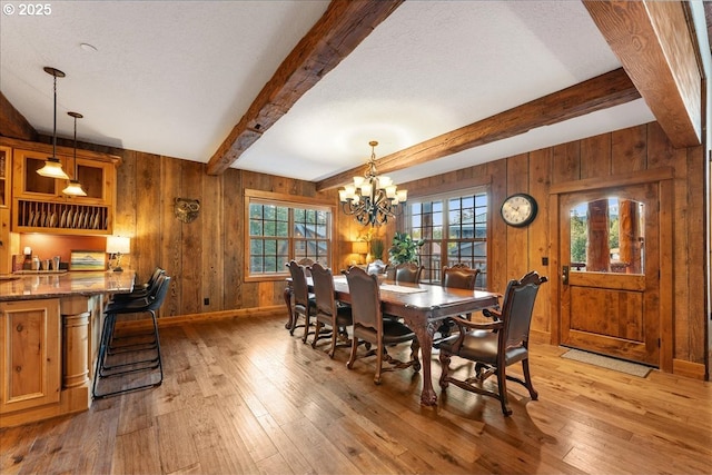 dining room featuring wood walls, light wood-style flooring, beamed ceiling, plenty of natural light, and a chandelier