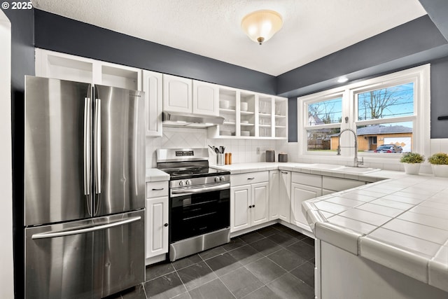 kitchen with a sink, stainless steel appliances, tile counters, under cabinet range hood, and white cabinetry