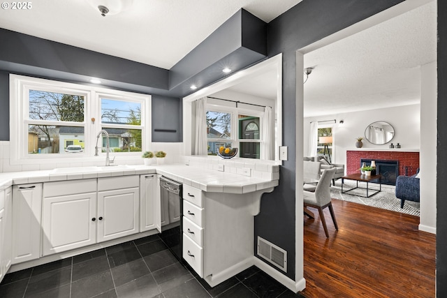 kitchen featuring visible vents, tile countertops, dishwashing machine, a brick fireplace, and a healthy amount of sunlight