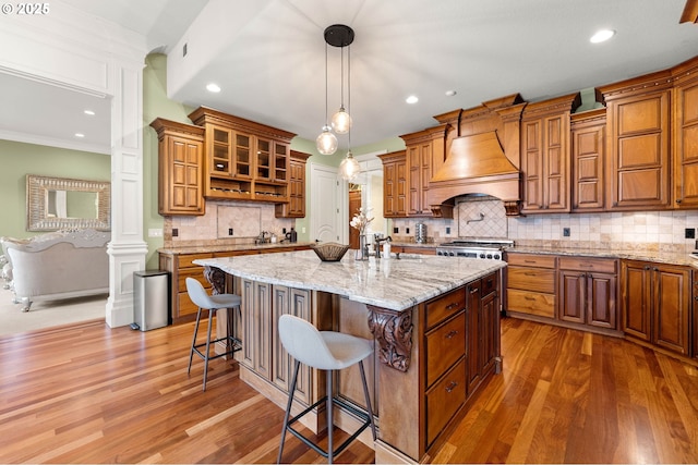 kitchen featuring premium range hood, a breakfast bar, an island with sink, pendant lighting, and light stone countertops