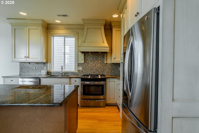 kitchen with sink, light hardwood / wood-style flooring, premium range hood, dark stone countertops, and stainless steel appliances