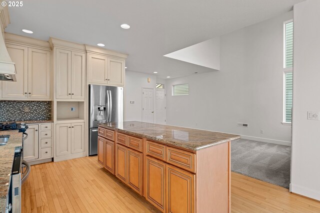 kitchen with light wood-type flooring, stainless steel fridge with ice dispenser, a kitchen island, cream cabinetry, and light stone countertops