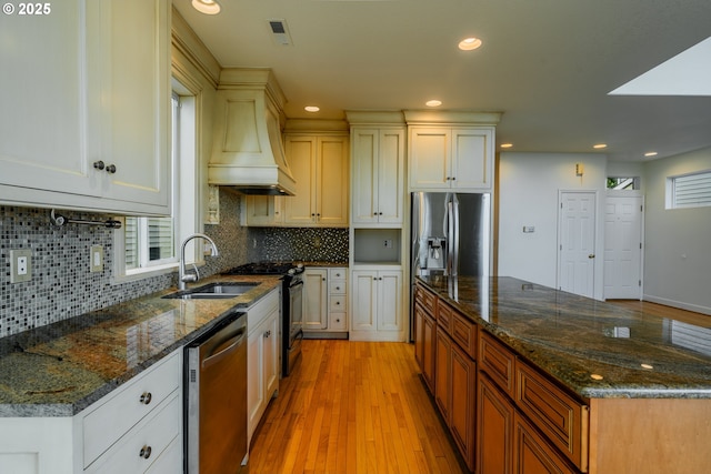 kitchen with sink, light hardwood / wood-style flooring, premium range hood, appliances with stainless steel finishes, and dark stone counters