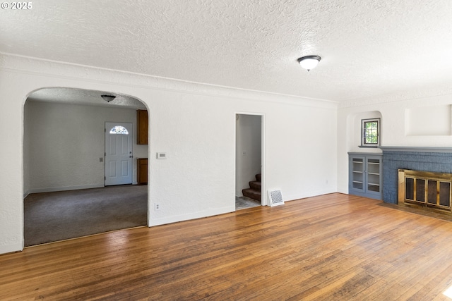 unfurnished living room with arched walkways, a textured ceiling, stairs, wood finished floors, and a brick fireplace