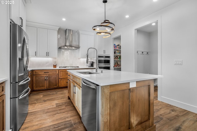 kitchen with wall chimney range hood, sink, appliances with stainless steel finishes, white cabinetry, and an island with sink