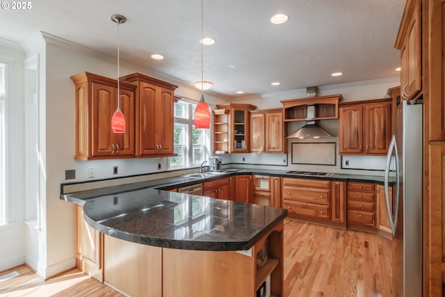 kitchen with wall chimney range hood, brown cabinetry, appliances with stainless steel finishes, open shelves, and a peninsula