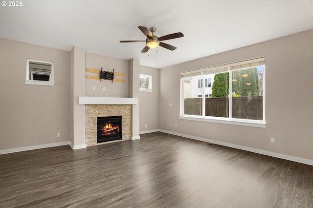 unfurnished living room with a stone fireplace, dark wood finished floors, a ceiling fan, and baseboards