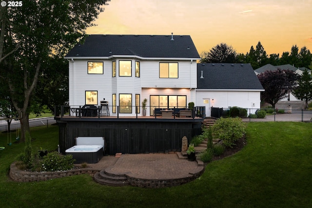 back of property at dusk with fence, a deck, a shingled roof, a hot tub, and a yard