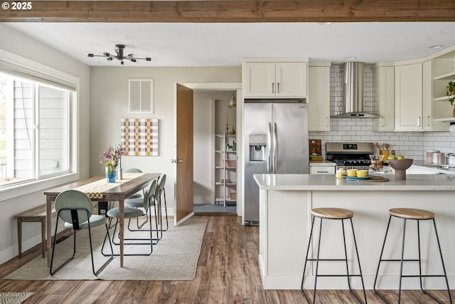 kitchen featuring wall chimney range hood, hardwood / wood-style floors, stainless steel appliances, tasteful backsplash, and a kitchen bar