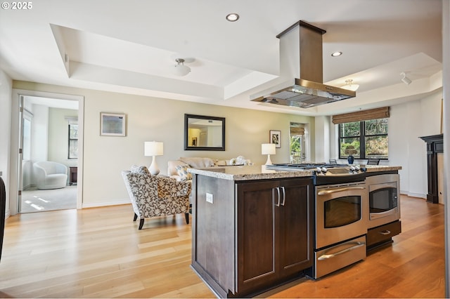 kitchen with stainless steel appliances, a raised ceiling, dark brown cabinetry, island range hood, and light wood-type flooring